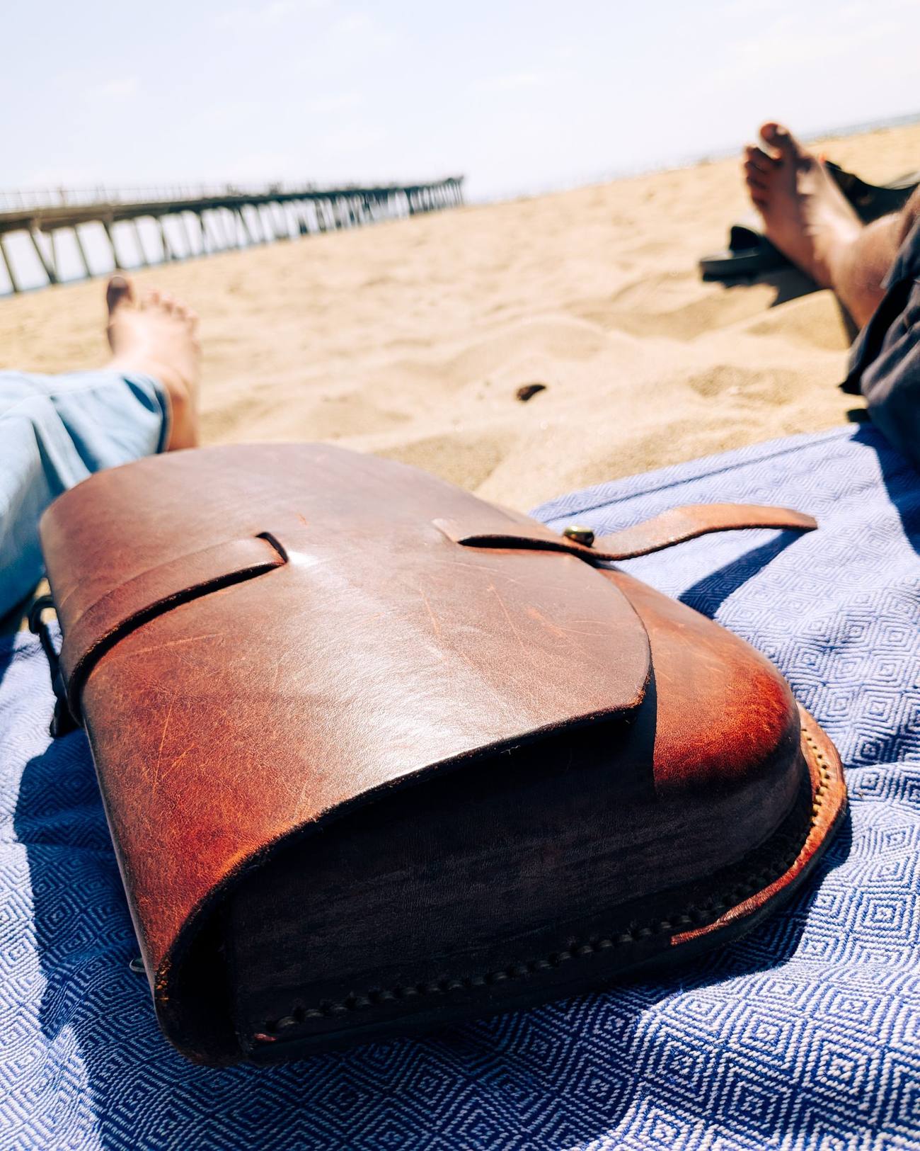 No. 0001, a chocolate wet-molded crossbody bag with a rich patina, resting on a beach blanket in the sun